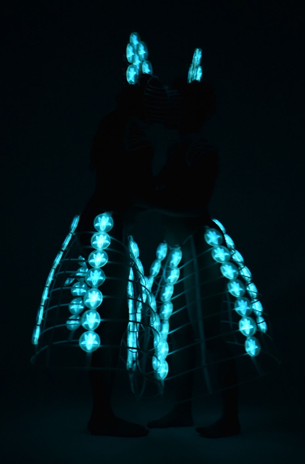 Embrace, figures illuminated by cyan bioluminescent forms in darkness